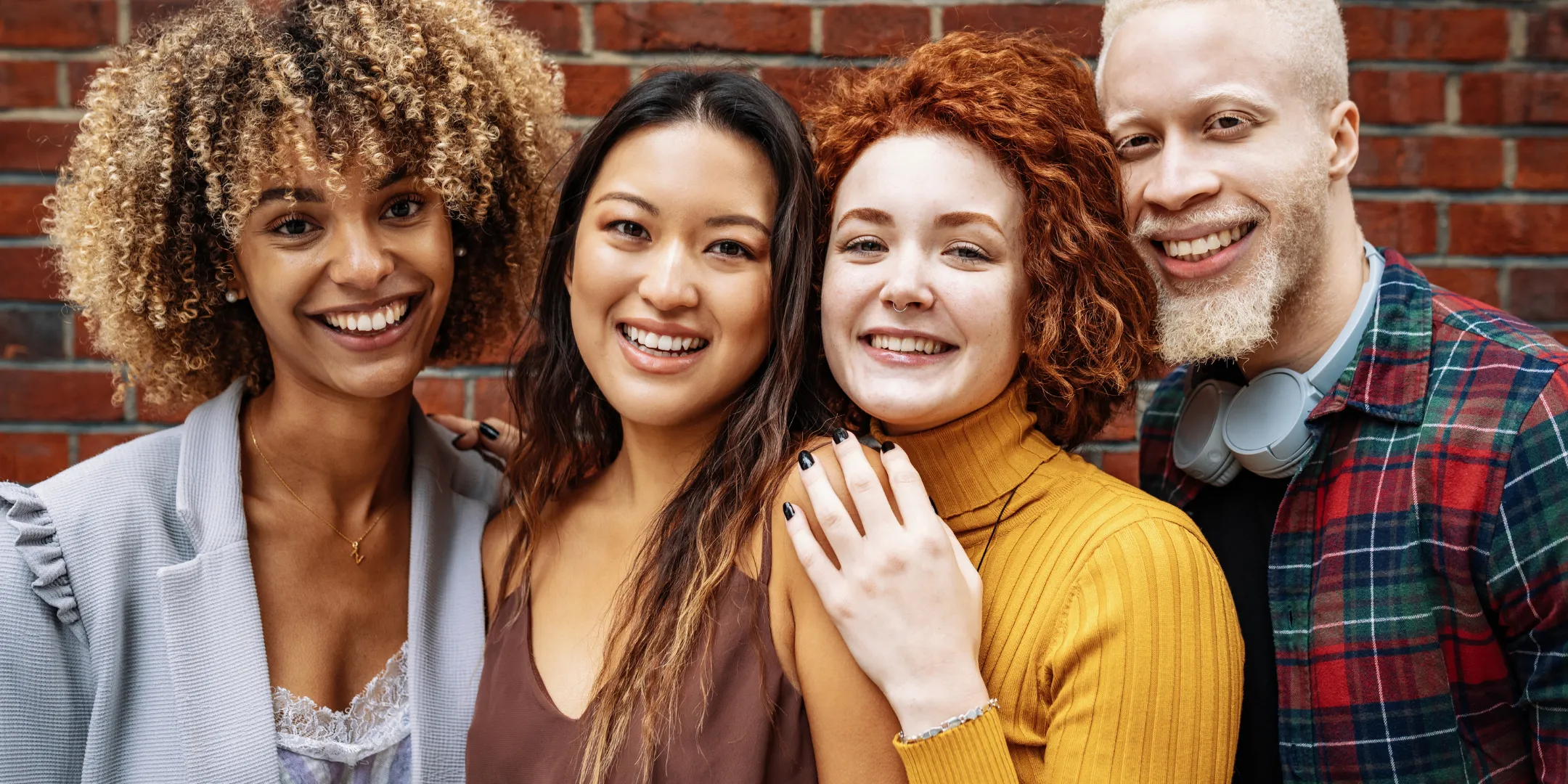 Four people stand close together in front of a brick wall, smiling warmly at the camera. The group is diverse in appearance, with different hair textures, skin tones, and styles, including curly, straight, and short light hair. They are dressed casually in layered tops, and one person has headphones resting around their neck. Their relaxed posture and closeness suggest friendship and a sense of connection.