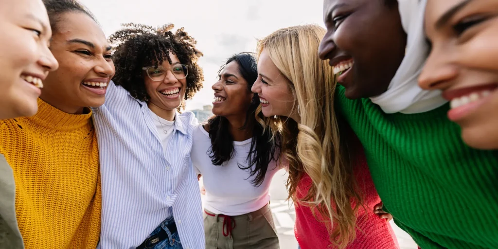 A diverse group of smiling young adults stand close together in a circle outdoors, laughing and looking at one another, conveying friendship, belonging, and connection.