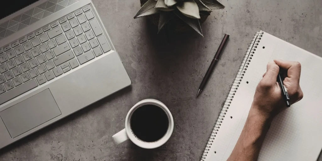This image shows a clean, minimal workspace viewed from above, with a focus on writing and thoughtful work.

On the left, part of a laptop is visible, suggesting digital work or research. In the centre, there’s a cup of black coffee, adding a sense of pause or rhythm to the workspace. Nearby, a pen rests on the surface, and a small plant brings in a subtle natural element.

On the right, a hand is actively writing in a spiral-bound notebook with grid paper. The posture of the hand and pen suggests focus and intention—capturing ideas, planning, or reflecting.

Overall, the scene feels calm and purposeful, blending productivity with a sense of quiet concentration and clarity.