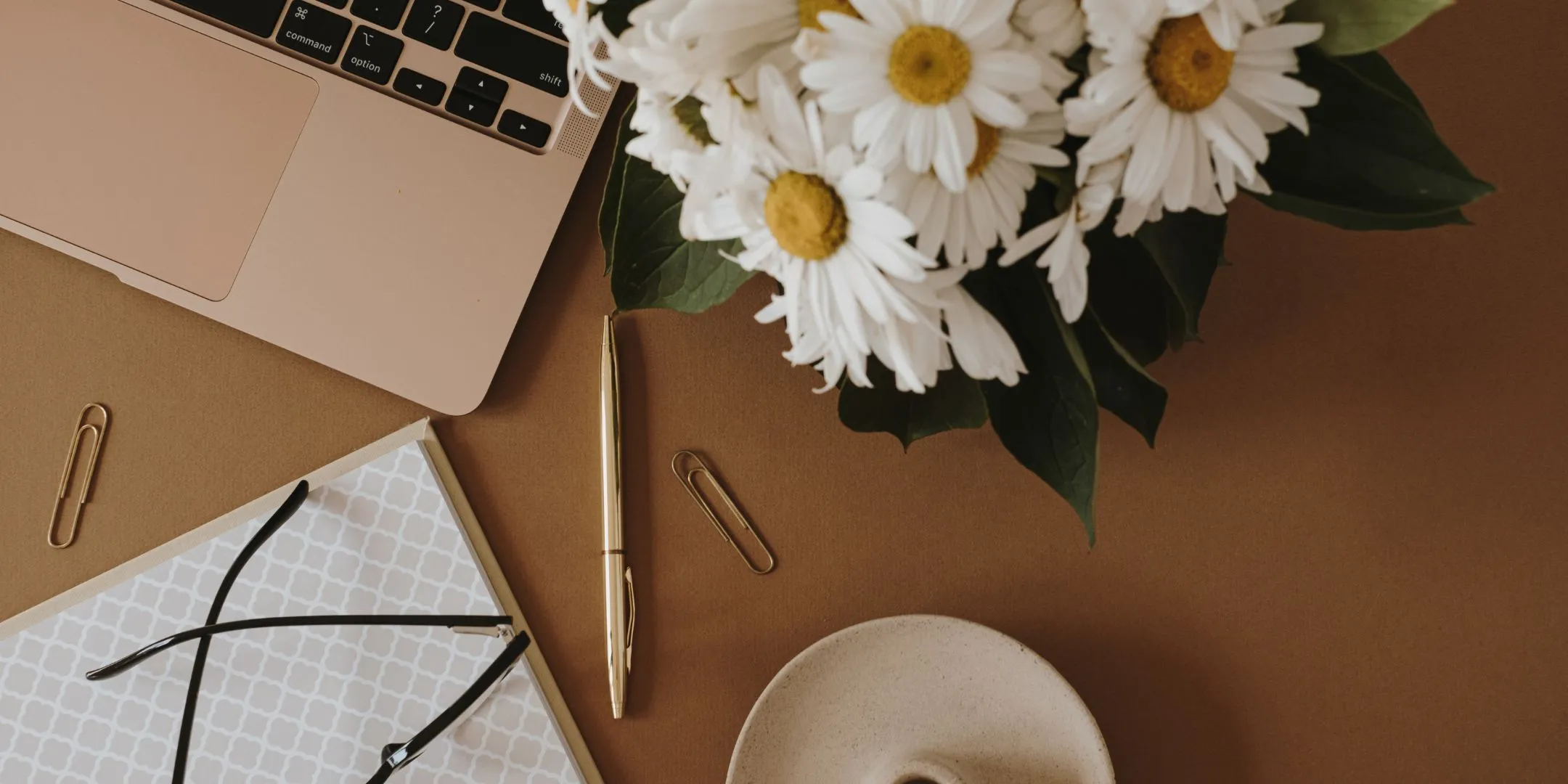 This image shows a neatly arranged workspace viewed from above, creating a calm and organised feel.  On the left, part of a laptop is visible, suggesting a work or study setting. Nearby, there’s a notebook with a subtle patterned cover, a pair of glasses resting on top, and a pen placed alongside it—items that hint at writing, planning, or reflection. A couple of paper clips are scattered lightly, adding to the everyday desk detail.  On the right, a bouquet of white daisies brings a soft, natural element into the scene, balancing the more structured objects with something gentle and grounding.  Overall, the image conveys a sense of quiet focus, organisation, and care—an environment that feels both productive and soothing.